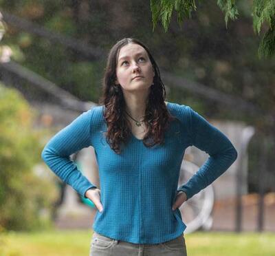 A person stands on campus path and looks up at the branches of an evergreen tree.