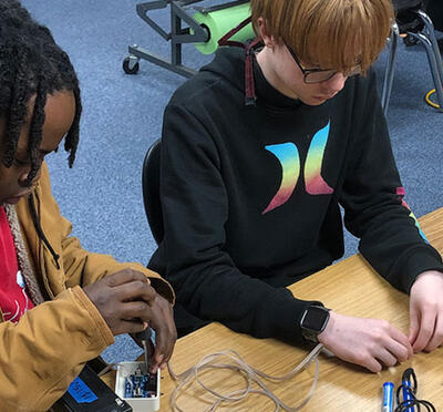 Two students sit at a table and adjust robotics equipment. 