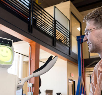 A person sits in a lab and looks at a robot with a smiling face and arms raised upward.