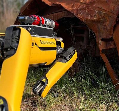 A robot with four legs steps over grass toward a rust covered vehicle.