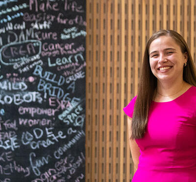 A person stands in front of a chalkboard with engineering terms written on it and smiles toward the camera.