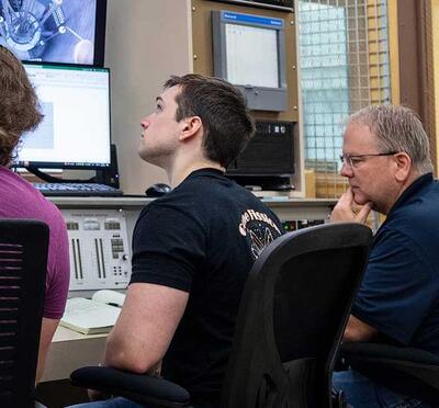 Three people sit in chairs and look at control systems of a nuclear reactor.