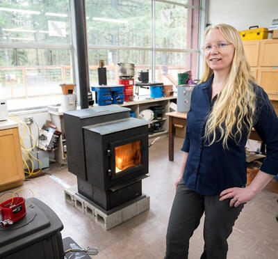 A woman with long blonde hair stands in a lab with a lit stove. Shelves of equipment and large windows displaying greenery are in the background.