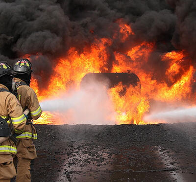 Six firefighters in protective clothing approach a burning tank.