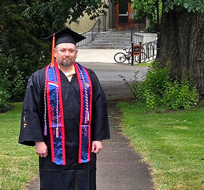 A person stands outside and looks toward the camera wearing academic regalia.