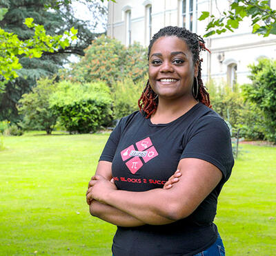 A person stands outside and smiles toward the camera with green grass and a historic building in the background.
