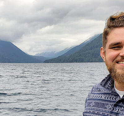 A person stands in front of a body of water with mountains in the distance and smiles toad the camera.