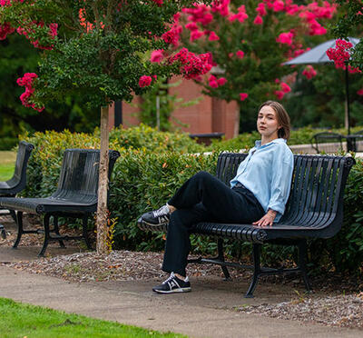 A person sits on a bench outside with summer blooms overhead and looks toward the camera.