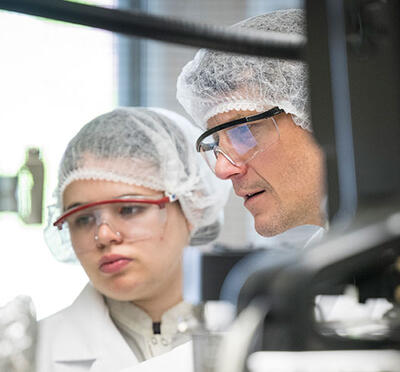 Two people wear lab coats and hair covers in a cleanroom and examine equipment.