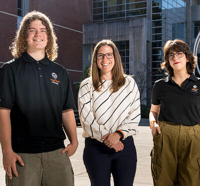 Three people stand outdoors on a campus with circular sculpture in the background and smile toward the camera. 