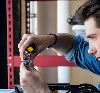 A person adjusts a small device with a tool and faces a window in a lab.