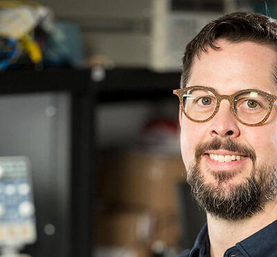 A person sits in a lab with equipment and digital screens in the background and smiles toward the camera.