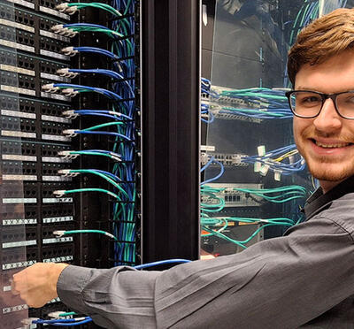 A person stands in a data center and adjusts hardware while looking toward the camera and smiling.