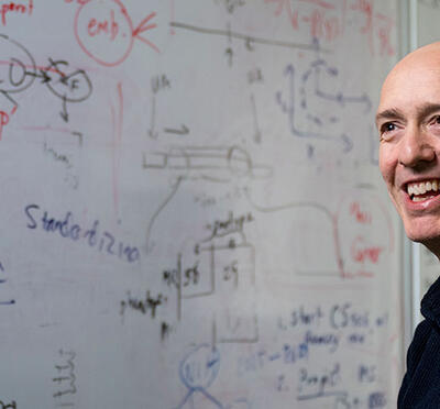 A person stands in front of a whiteboard with figures on it and smiles toward a subject of camera.