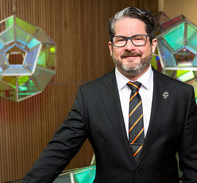 A person in a suit stands near a railing with a glass art installation behind.