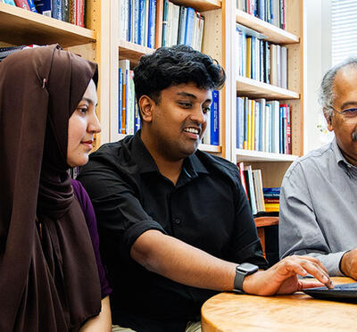 Three people sit at a table in an office and look at a laptop. 