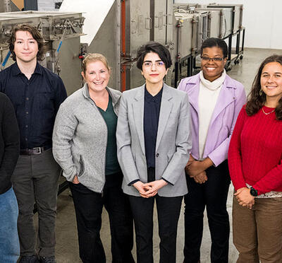 Seven people stand in a lab next to cylindrical equipment and look up toward the camera.