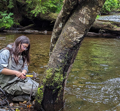 Two women install sensing equipment in a stream. The scene includes clear flowing water, moss-covered rocks, and fallen logs in the background.