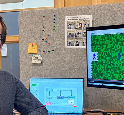 Person seated at a desk with two monitors; the larger monitor displays a green grid-based game with colored characters, while the smaller laptop screen shows a flowchart diagram.