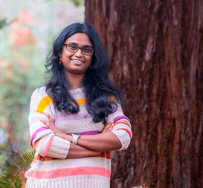 A person wears a striped sweater and stands outside in front of a large tree with green vegetation in the foreground.