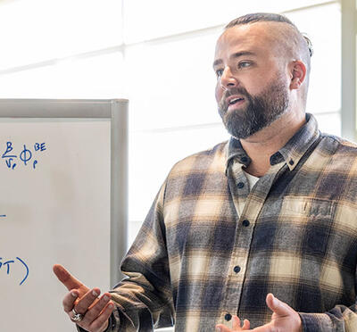 A person wears a plaid shirt and stands indoors near a whiteboard with complex mathematical equations.