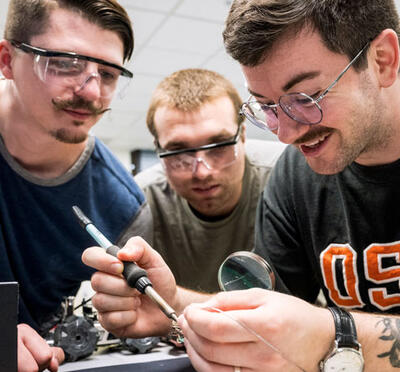 Three men wearing safety goggles collaborate on an electronics project. They appear focused and engaged, with one holding a soldering iron.