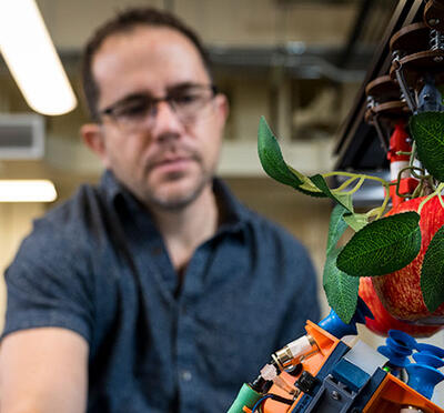 A person in a lab setting looks down at a robotic arm next to an apple. 