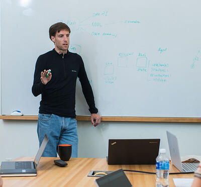 A person stands at the front of a conference room, pointing to a whiteboard covered with handwritten diagrams and notes, while several people sit around a table with laptops and notebooks, listening and discussing.