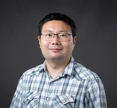 A person wears a light plaid button-up shirt and stands against a plain dark background, facing the camera.