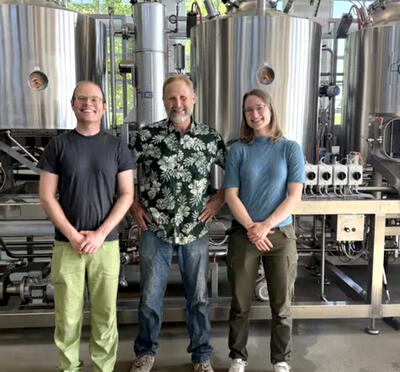 Three people standing in front of brewery equipment.