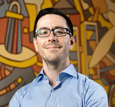 A person wears a light blue button‑down shirt and stands in front of a large, colorful mural featuring abstract shapes and industrial-themed illustrations.