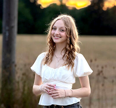 A person with shoulder-length hair stands in a field at sunset and wears a white short-sleeve shirt. 