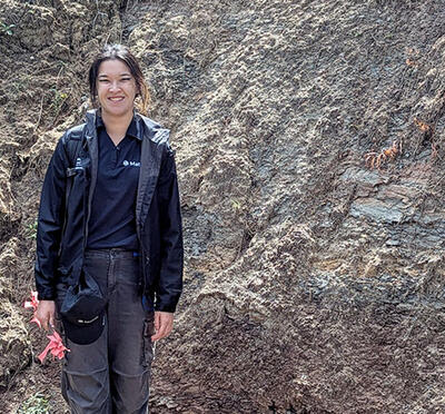 A person stands outdoors beside an exposed rock wall with sparse vegetation and small trees around them.
