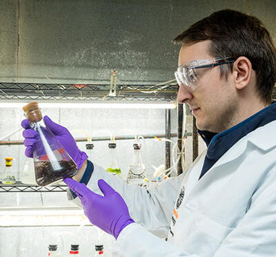 A person in a lab wears a lab coat and gloves while holding and examining a glass flask containing a dark liquid, standing in front of metal shelving.