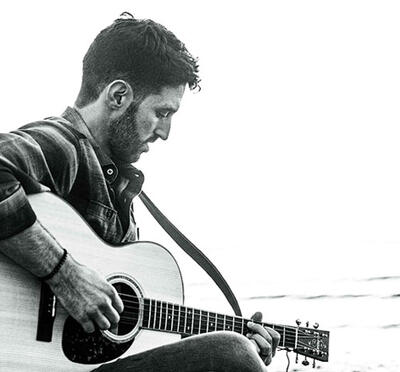 A person sits on a beach, playing an acoustic guitar with ocean waves and distant rocks in the background.
