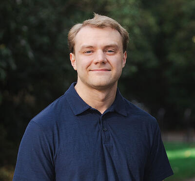 A person stands on a paved path with green grass and trees and looks toward the camera.