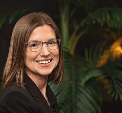 Camille Palmer wears business attire and sits in a dark area with tropical vegetation in the background and smiles toward the camera. 