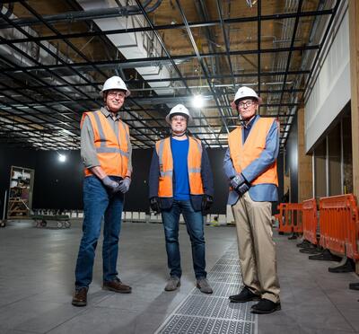 Anthony Koppers, Shayne Huddleston, and Todd Palmer dressed in hard hats and safety vests in a dark room under construction.
