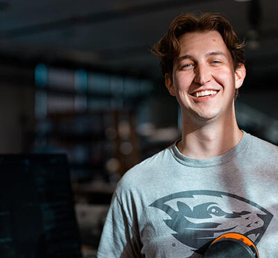 Alexander Roller wears a grey Oregon State Beavers t-shirt and stands in a workspace with sunlight illuminating his face and the background in dark shadow. 