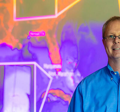 A person in business attire stands in a classroom with a digital projection of colorful bathymetry images behind them.