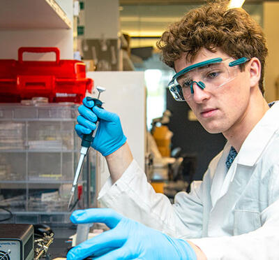 A man in a white lab coat sits at a lab bench, handling equipment and taking notes.