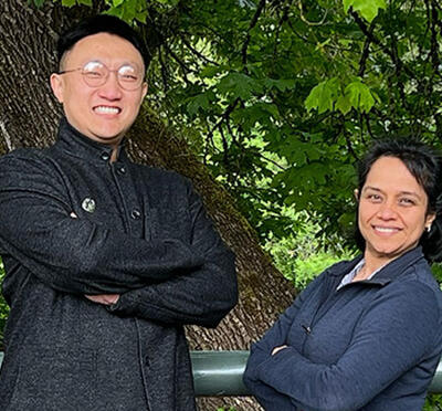Two people wear black and stand outside in front of vegetation and smile toward the camera.