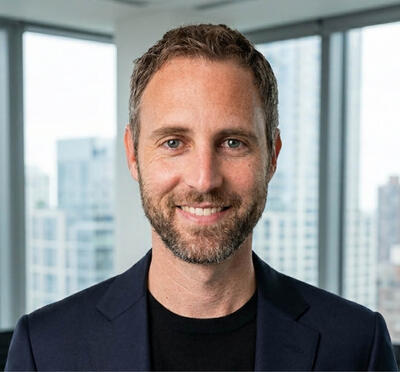 A person wears a dark suit in an office setting and looks toward the camera with tall windows and a city skyline in the background.