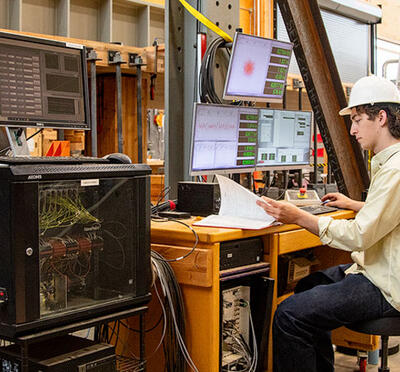 Dylan Goff wears a hard hat and sits in front of a computer and monitoring equipment in the Emmerson Lab, a large timber engineering facility.