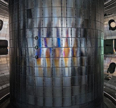 A person wears a clean-room suit with hair covering, gloves, and boot covers and stands inside a large cylindrical chamber lined with metallic panels and equipment.