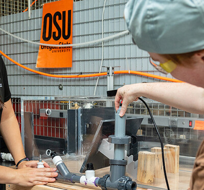 Kathryn Newhart and Perry Wilson wear eye protection and stand next to a workbench in a lab and adjust a device with two intersecting pipes.