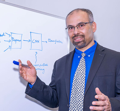 Saurabh Bagchi wears a blue suit and stands in front of a whiteboard with a hand-drawn illustration of workflow on the board behind him.
