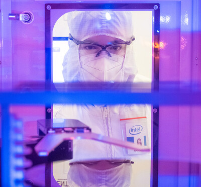 A person wears clean-room protective clothing, safety glasses, and a face mask is viewed through the interior of semiconductor manufacturing equipment in purple and blue light. 