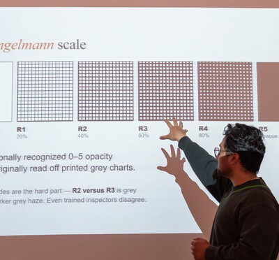 A person points to a projected image of the Ringelmann scale, showing a progression of shaded grids, from clear to opaque.