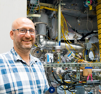 Guillaume Mignot stands outside beside a large instrument filled with pipes, cables, valves, and metal components.